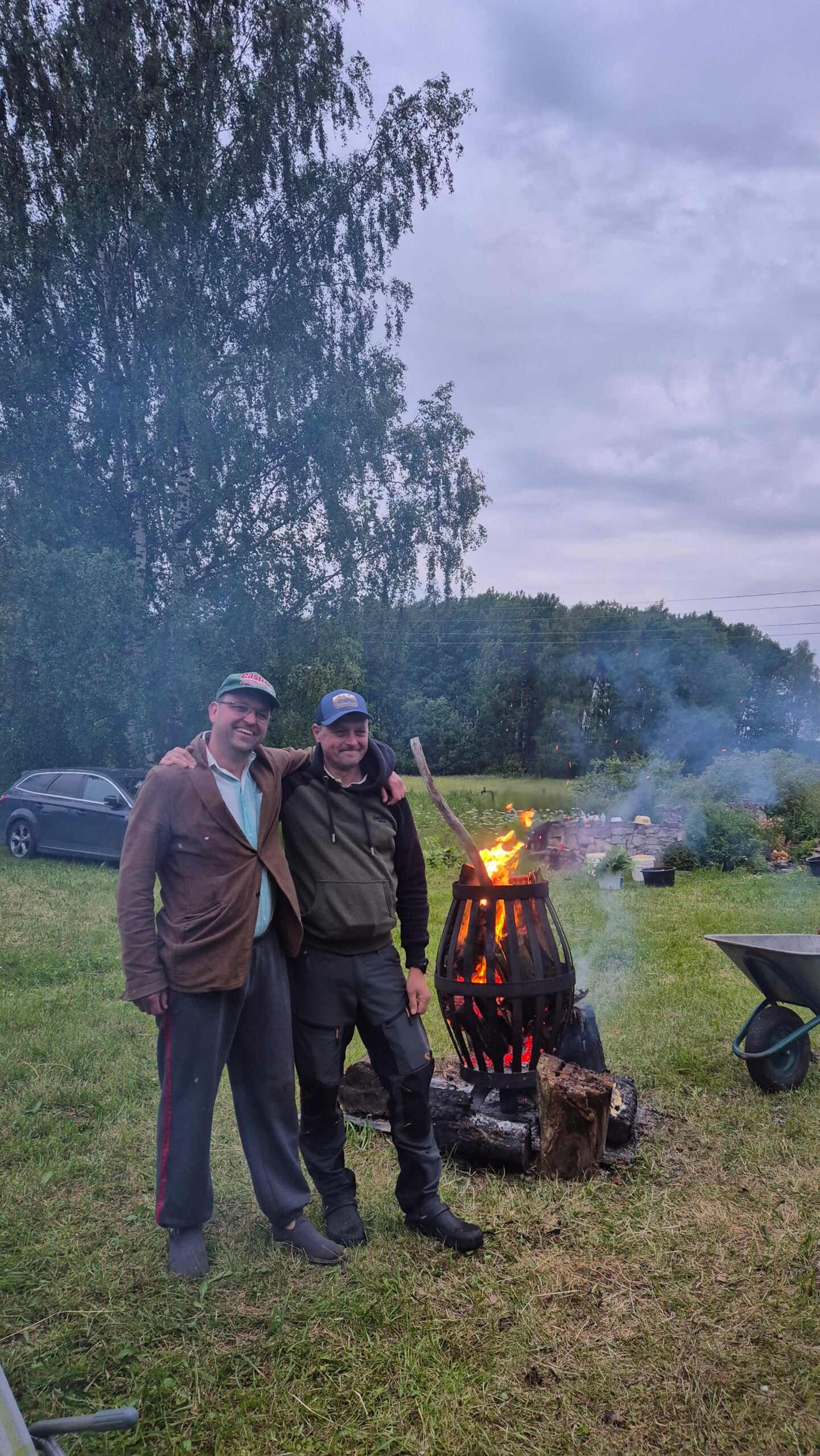 Jānis and a friend beside an SJR fire basket in the garden at dusk